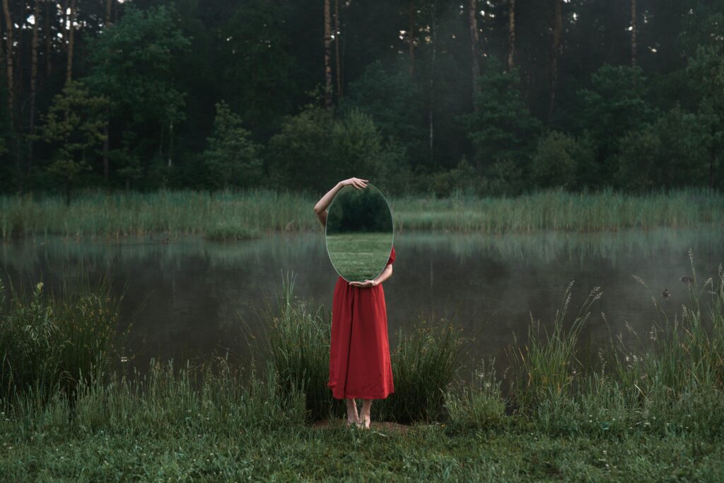 La ilusión del control: el ego frente a la incertidumbre Artistic photo of a woman in nature holding a mirror beside a misty pond.