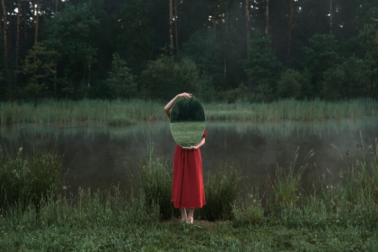 Artistic photo of a woman in nature holding a mirror beside a misty pond.